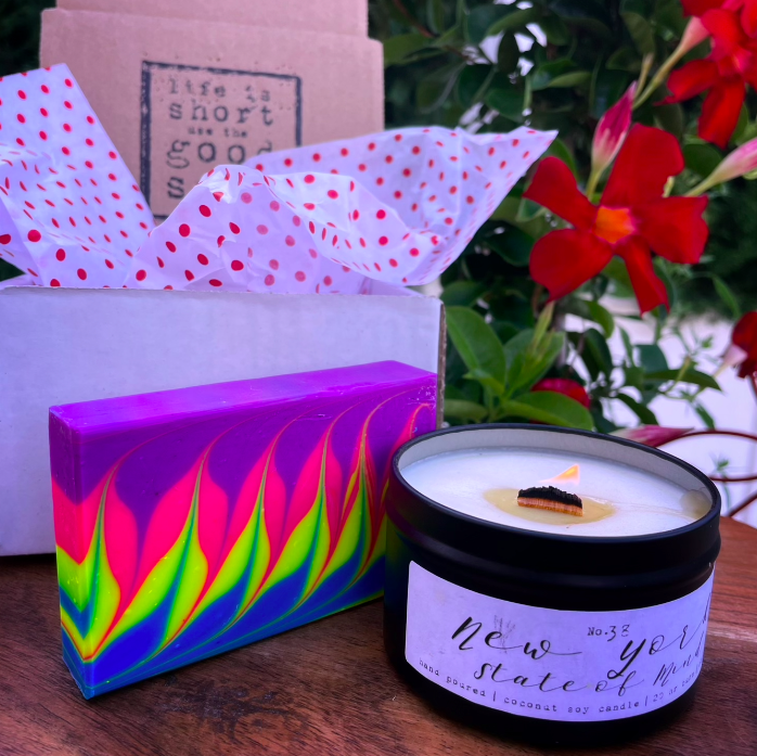 a colorful soap next to a candle in a black tin in front of a box and some flowers